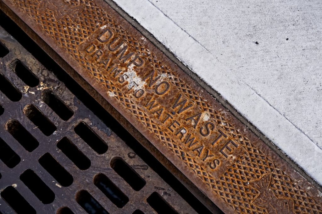 Close-up of a rusty drainage grate with environmental warning text on concrete sidewalk